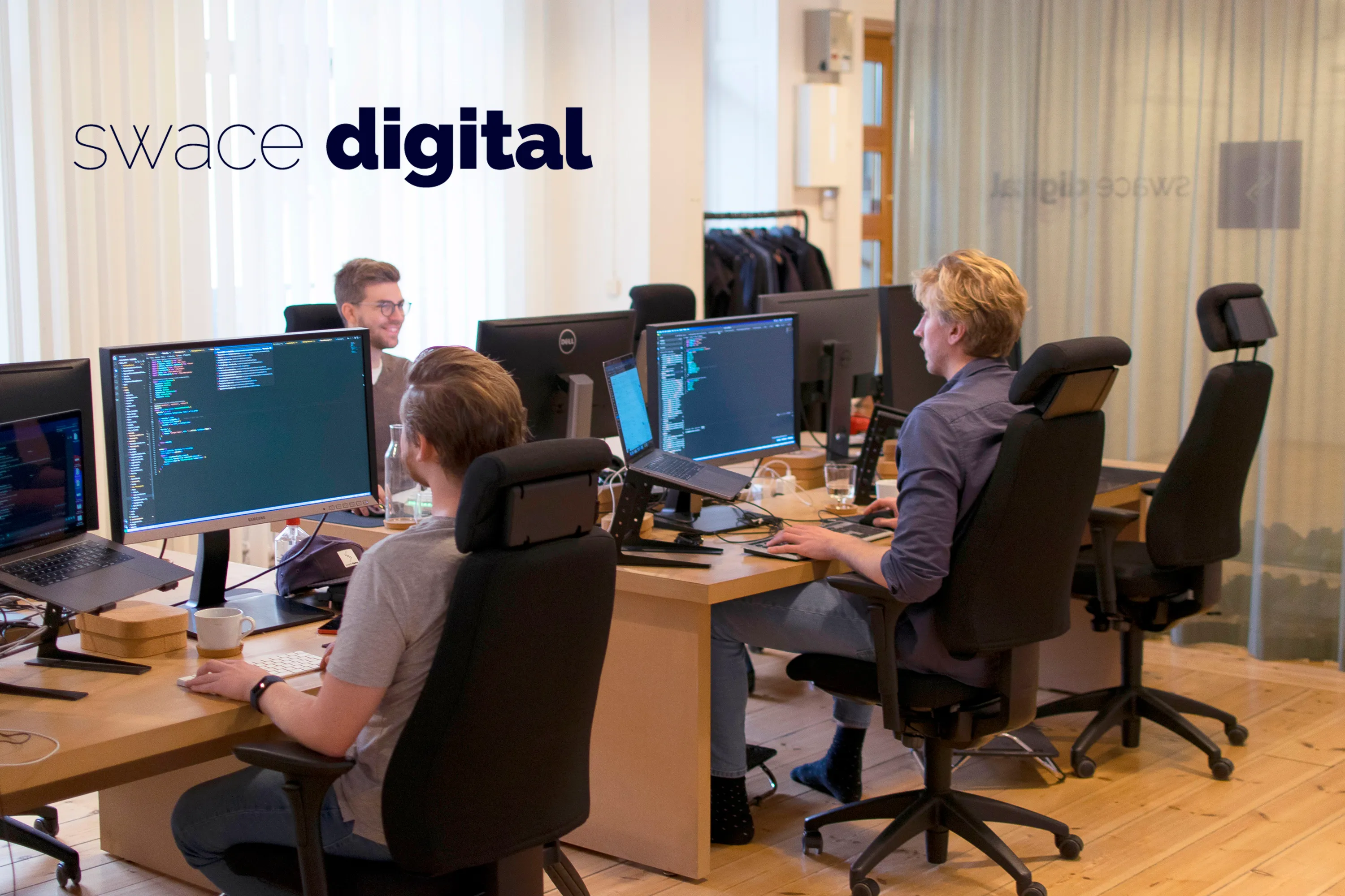 Tre personer som sitter framför datorskärmar och kodar på ett kontor.
