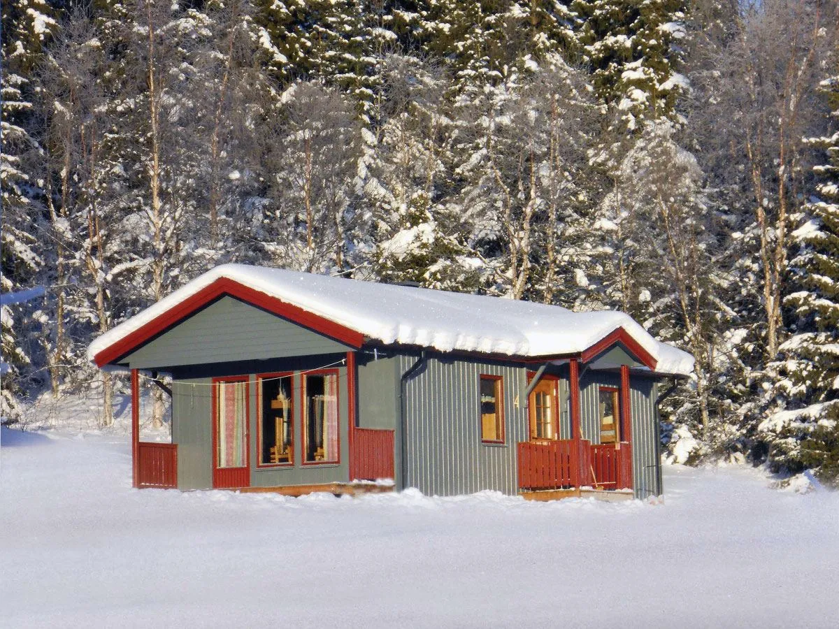 Stuga med snö på taket och skog bakom.